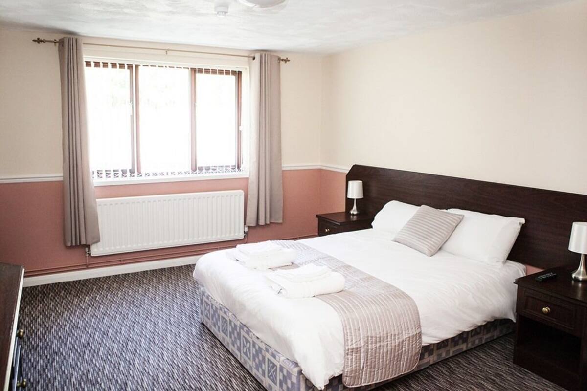 A hotel room with a large bed, a nightstand, a window with curtains, and a radiator.
