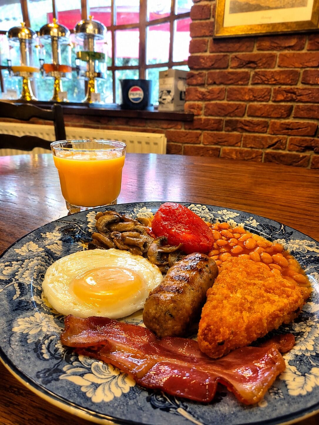 Une assiette de petit-déjeuner comprenant des œufs, du bacon, des saucisses, des haricots et des toasts, accompagnée d’un verre de jus d’orange.