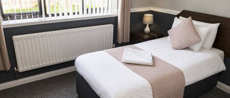 Una cama bien tendida con ropa blanca y una manta color beige, una lámpara de noche y una ventana con persianas.
