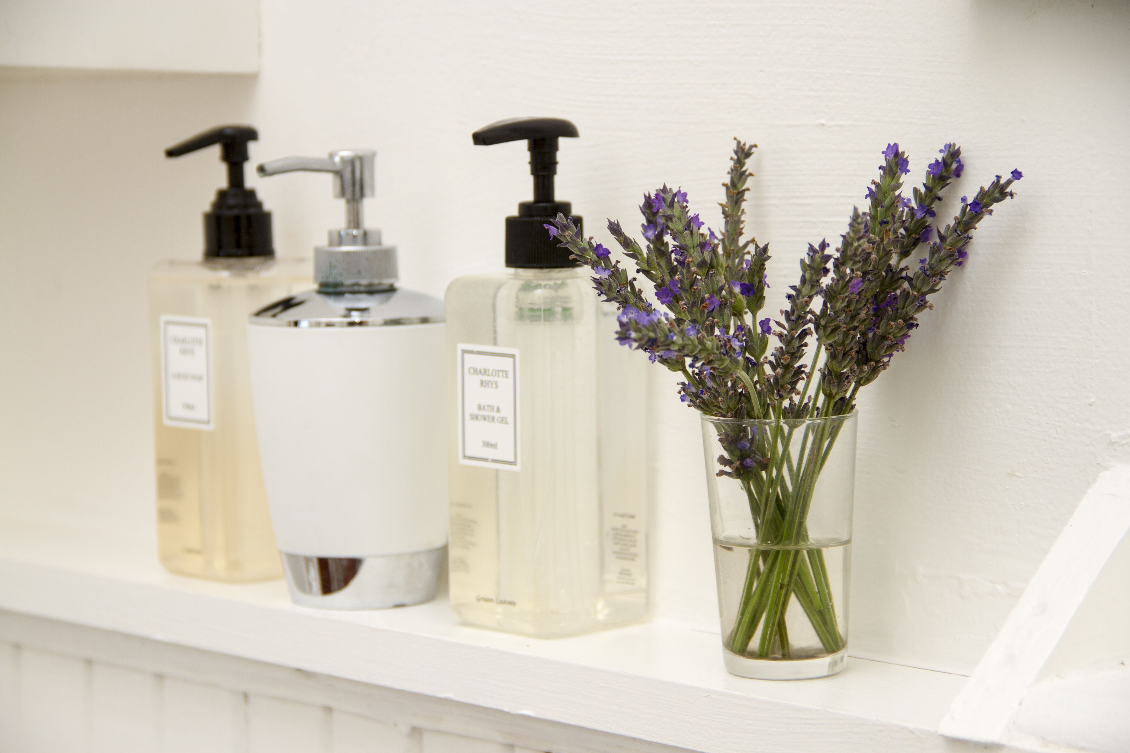 A shelf with three bottles of liquid soap and a glass vase with lavender flowers.