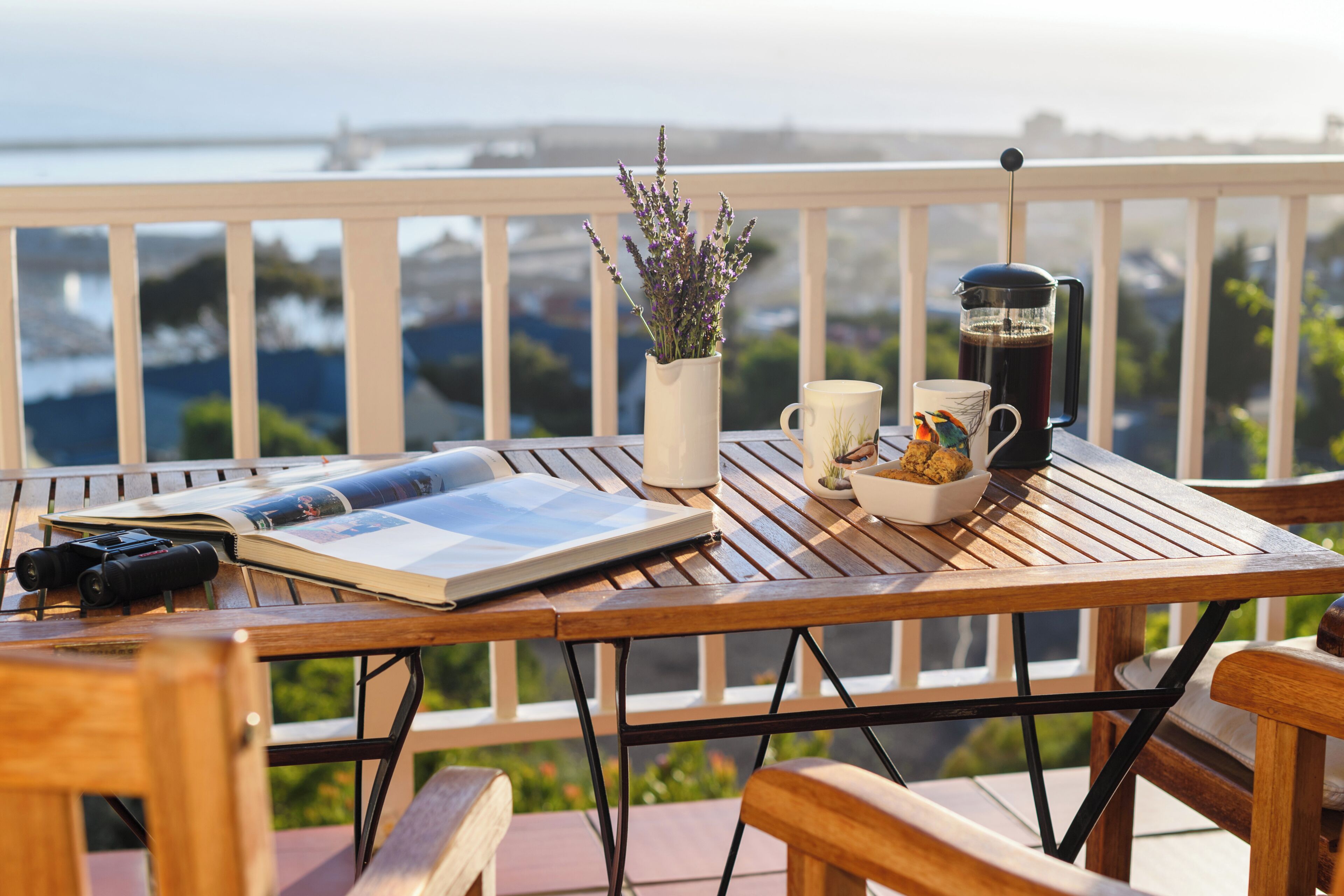 A wooden table with a book, a camera, a coffee pot, a mug, and a vase of flowers on a balcony overlooking a cityscape.