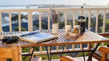 A wooden table with a book, a camera, a coffee pot, a mug, and a vase of flowers on a balcony overlooking a cityscape.