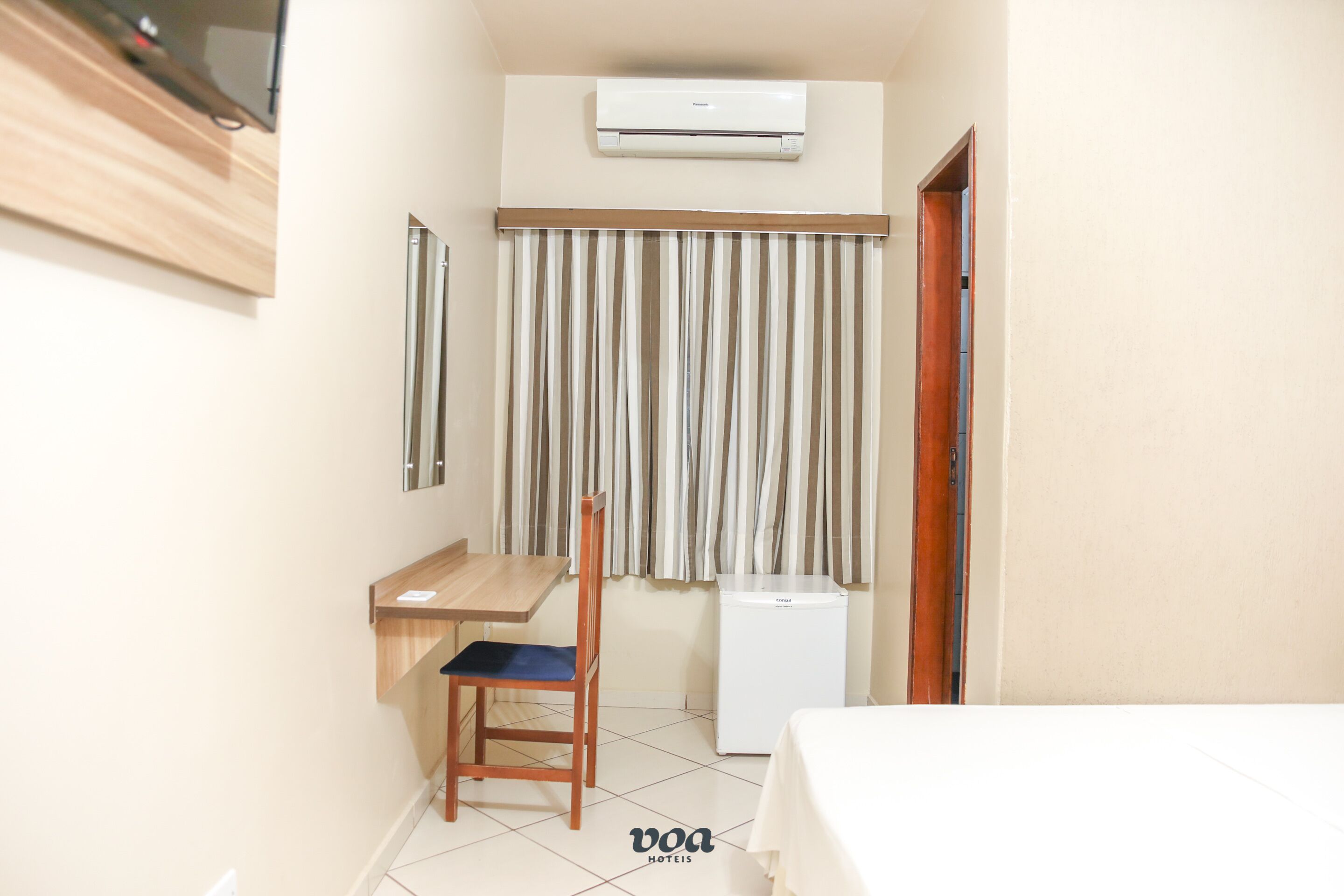 A hotel room with a desk, chair, bed, air conditioning unit, and curtains.