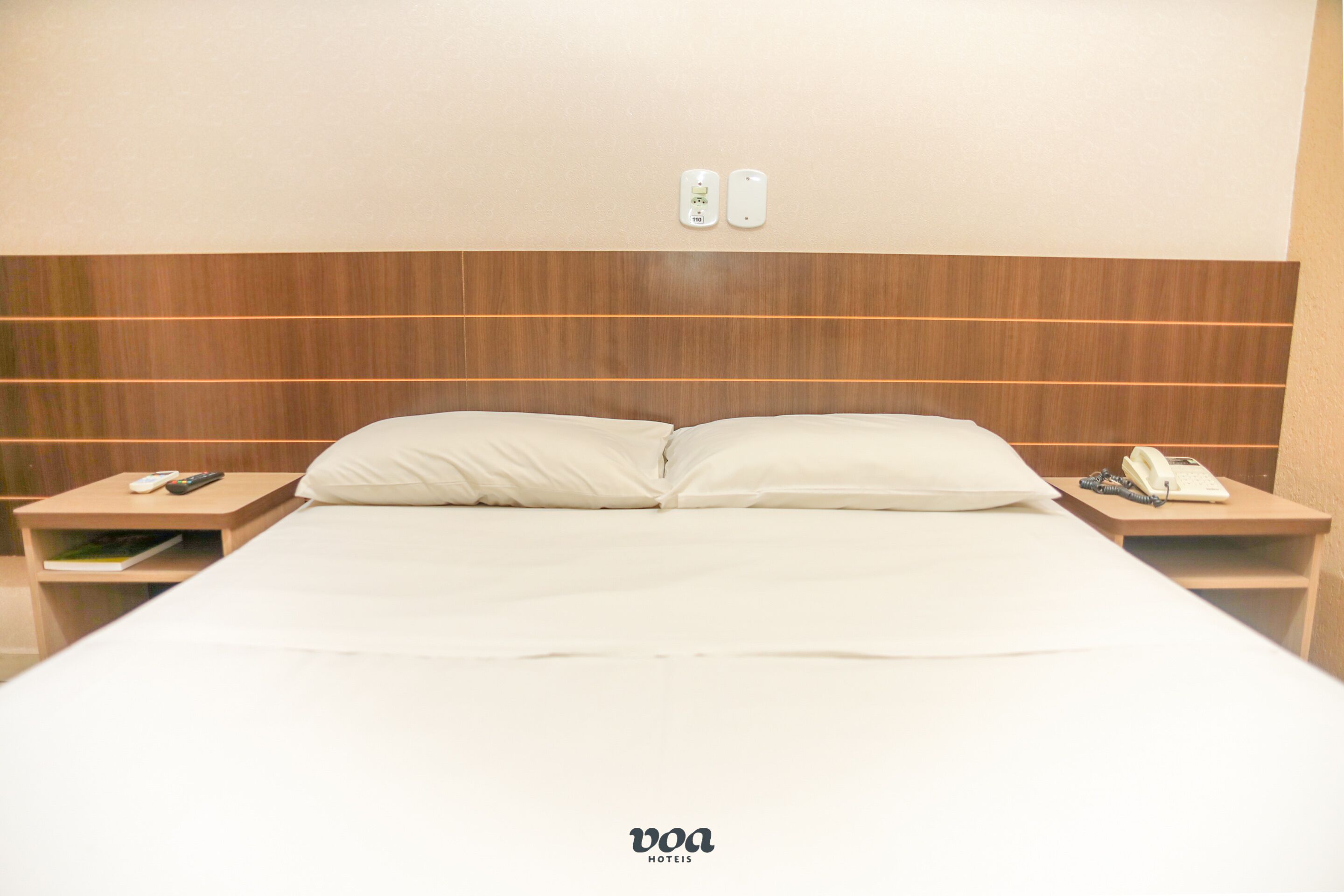 Ein Hotelzimmer mit einem hölzernen Kopfteil, einem Bett mit weißen Leinentüchern, Nachttischen mit Fernbedienung und Telefon sowie einer Minibar.