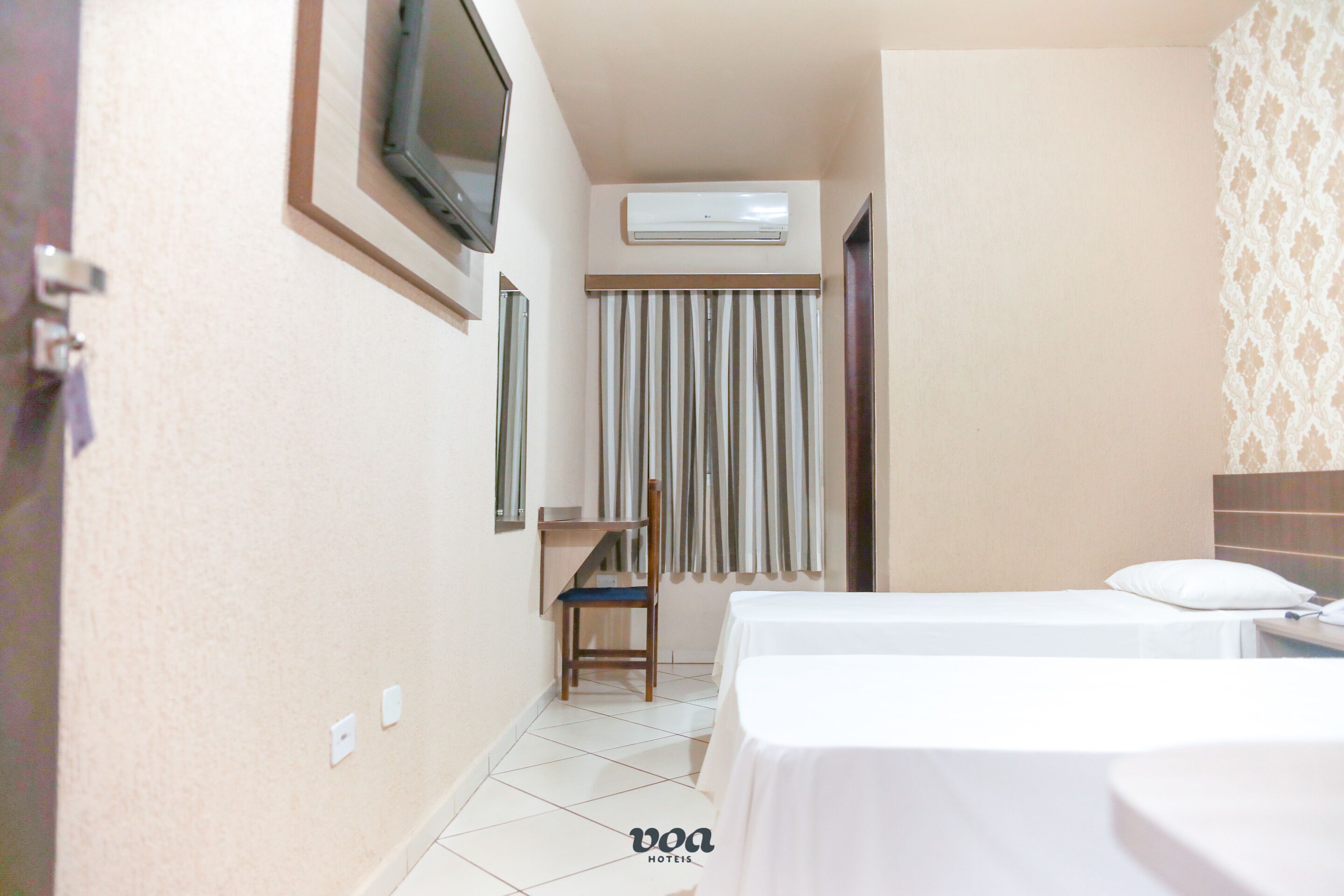 A hotel room with two beds, a TV mounted on the wall, an air conditioning unit, and a small desk with a chair.