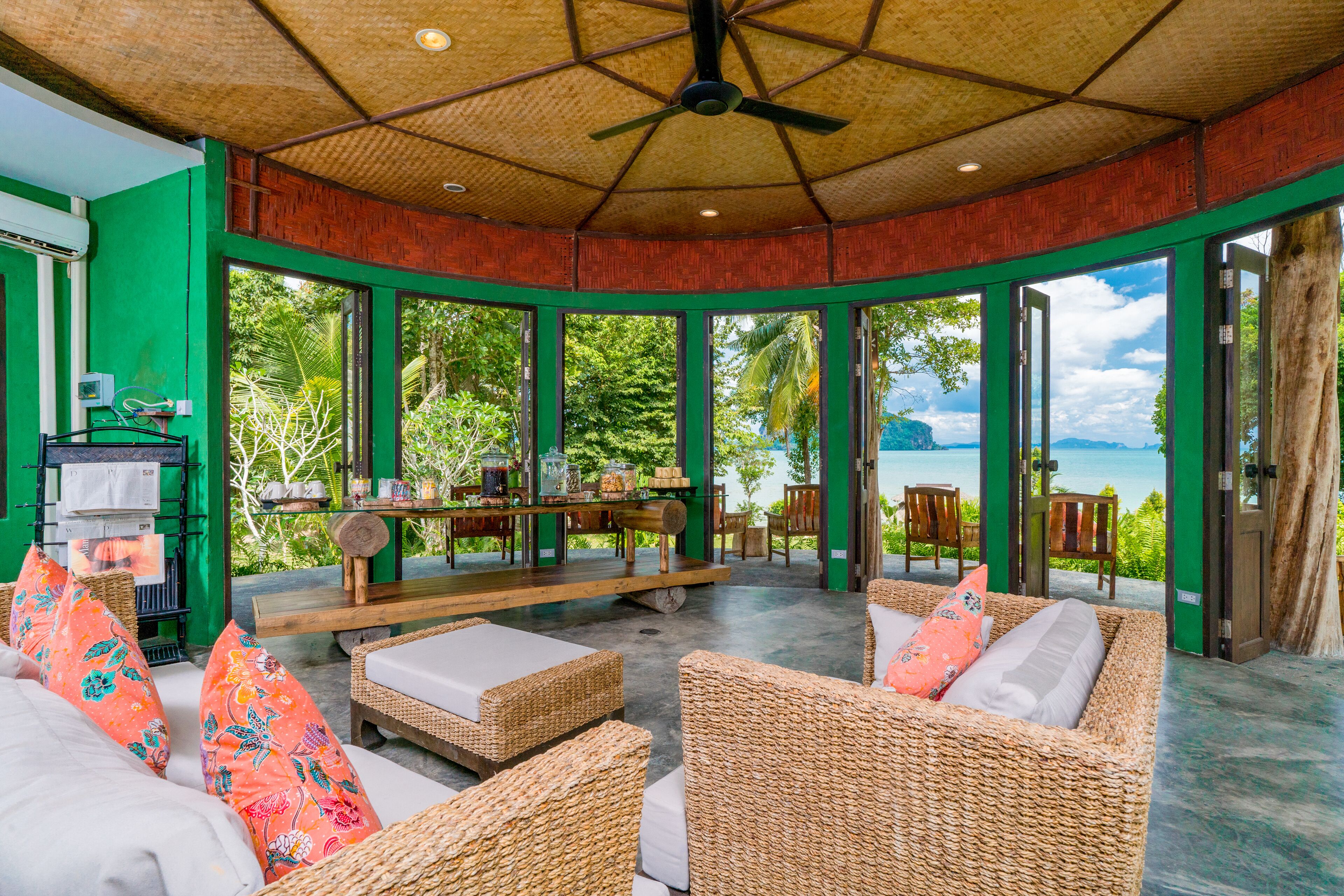 A spacious outdoor seating area with wicker furniture and a ceiling fan, overlooking a scenic view of the sea and lush greenery.