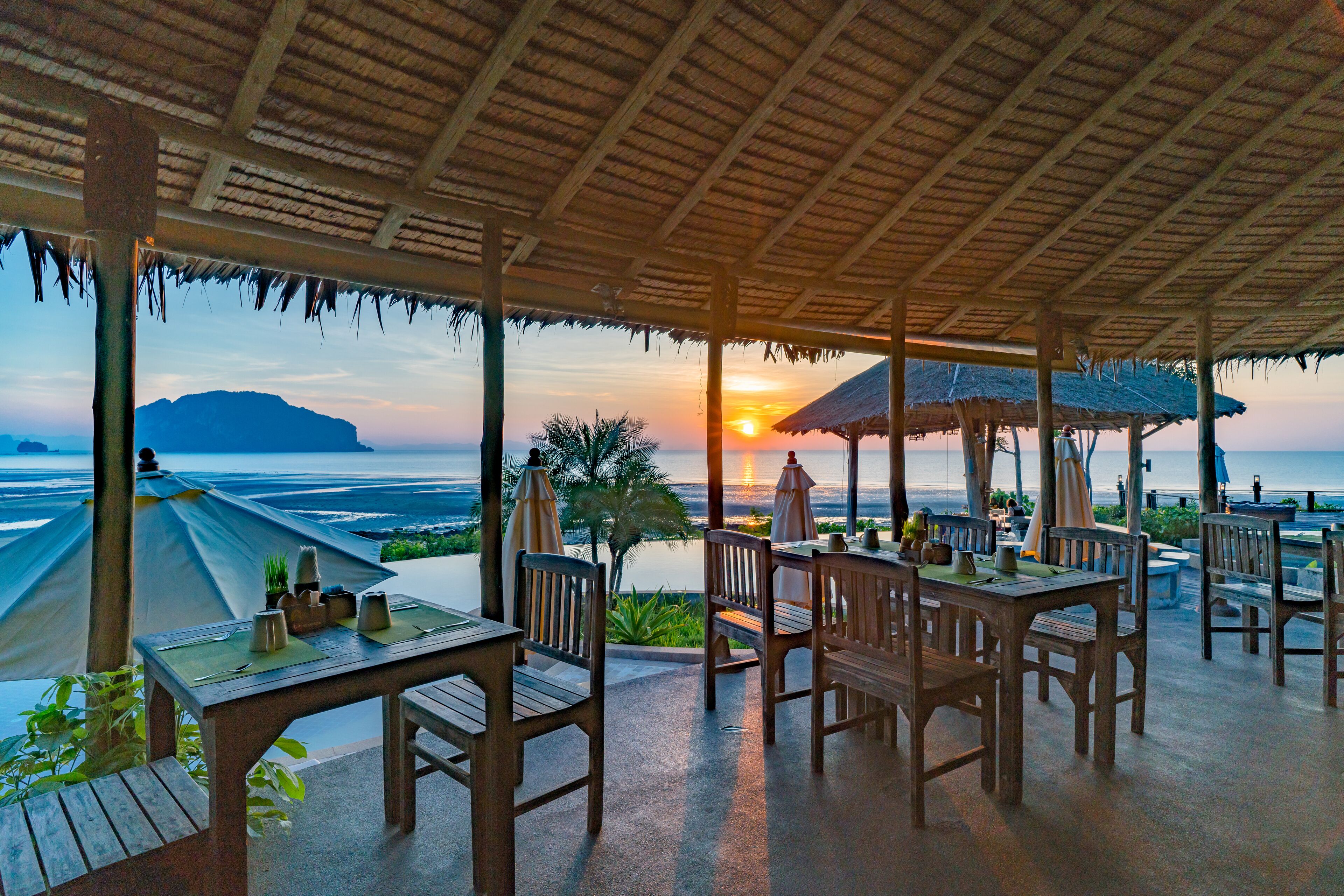 Un espace extérieur aménagé pour les repas, avec des tables et des chaises en bois, des parasols et une vue sur le coucher de soleil sur l’océan.
