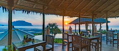 Un espace extérieur aménagé pour les repas, avec des tables et des chaises en bois, des parasols et une vue sur le coucher de soleil sur l’océan.