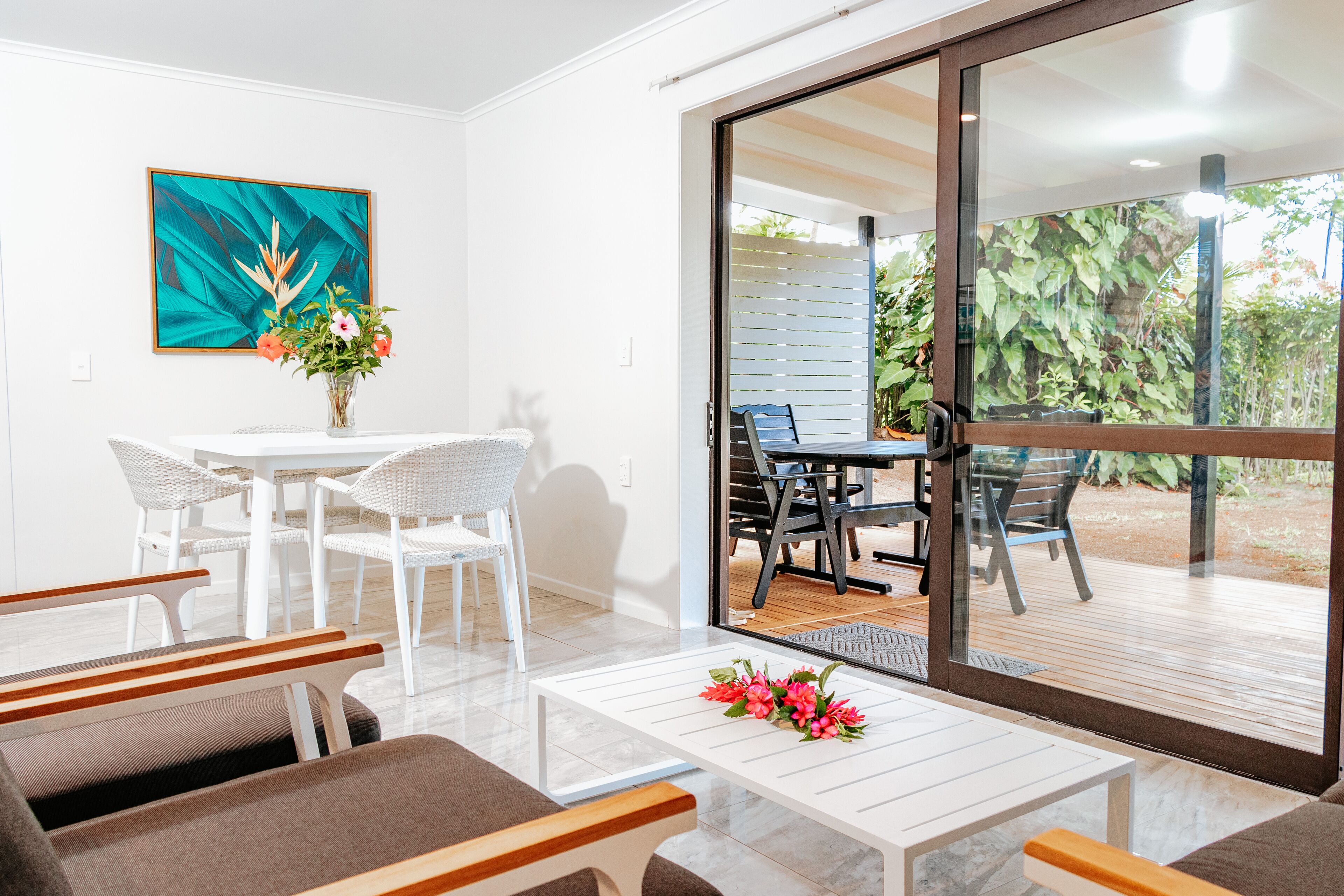 A modern living room with a white sofa, a coffee table with flowers, a dining area with a white table and wicker chairs, and a sliding glass door leading to an outdoor patio with a wooden deck and greenery.