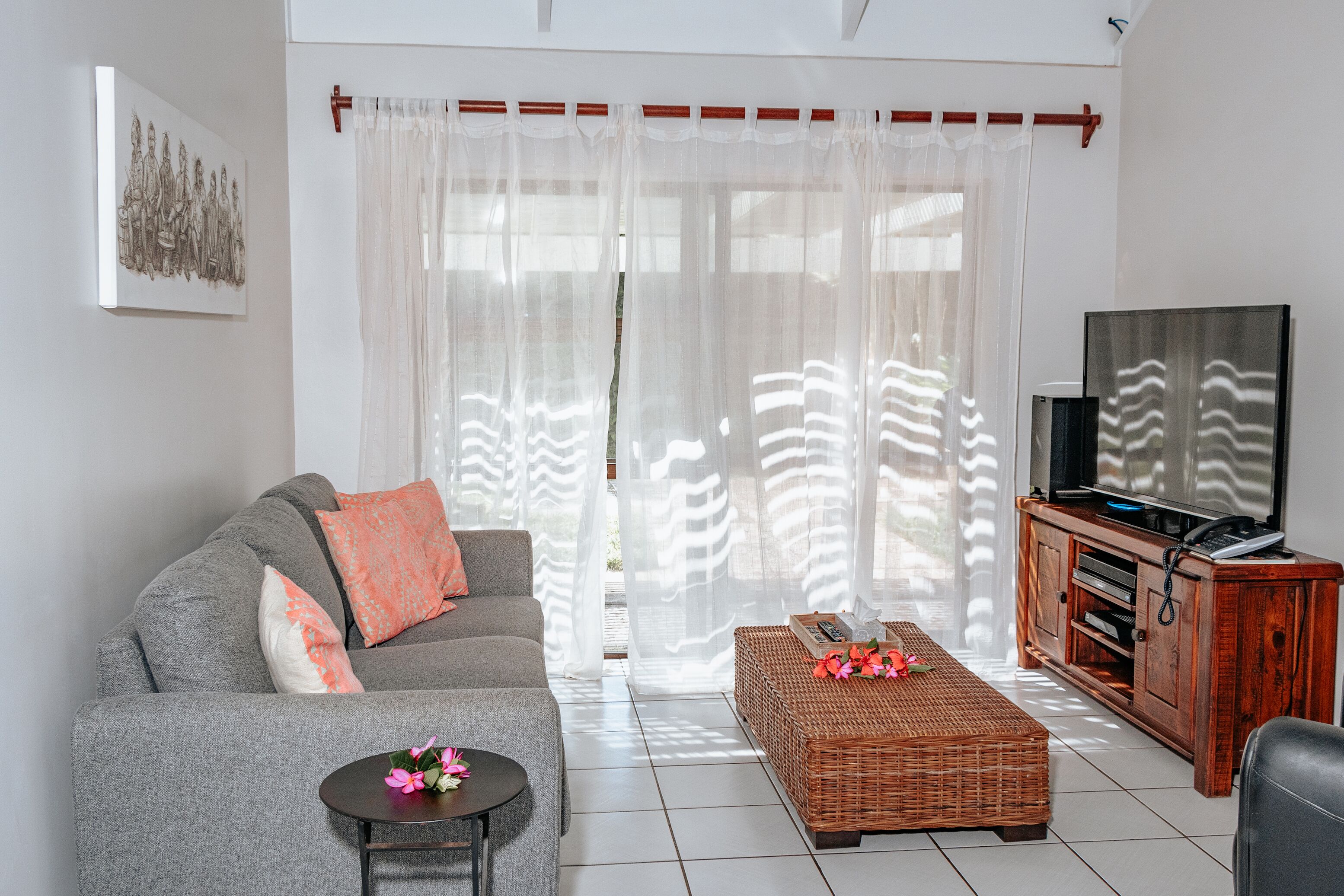 Een woonkamer met een grijze bank, een kleine ronde tafel met bloemen, een rieten poef, een televisie op een houten kast en dunne gordijnen.