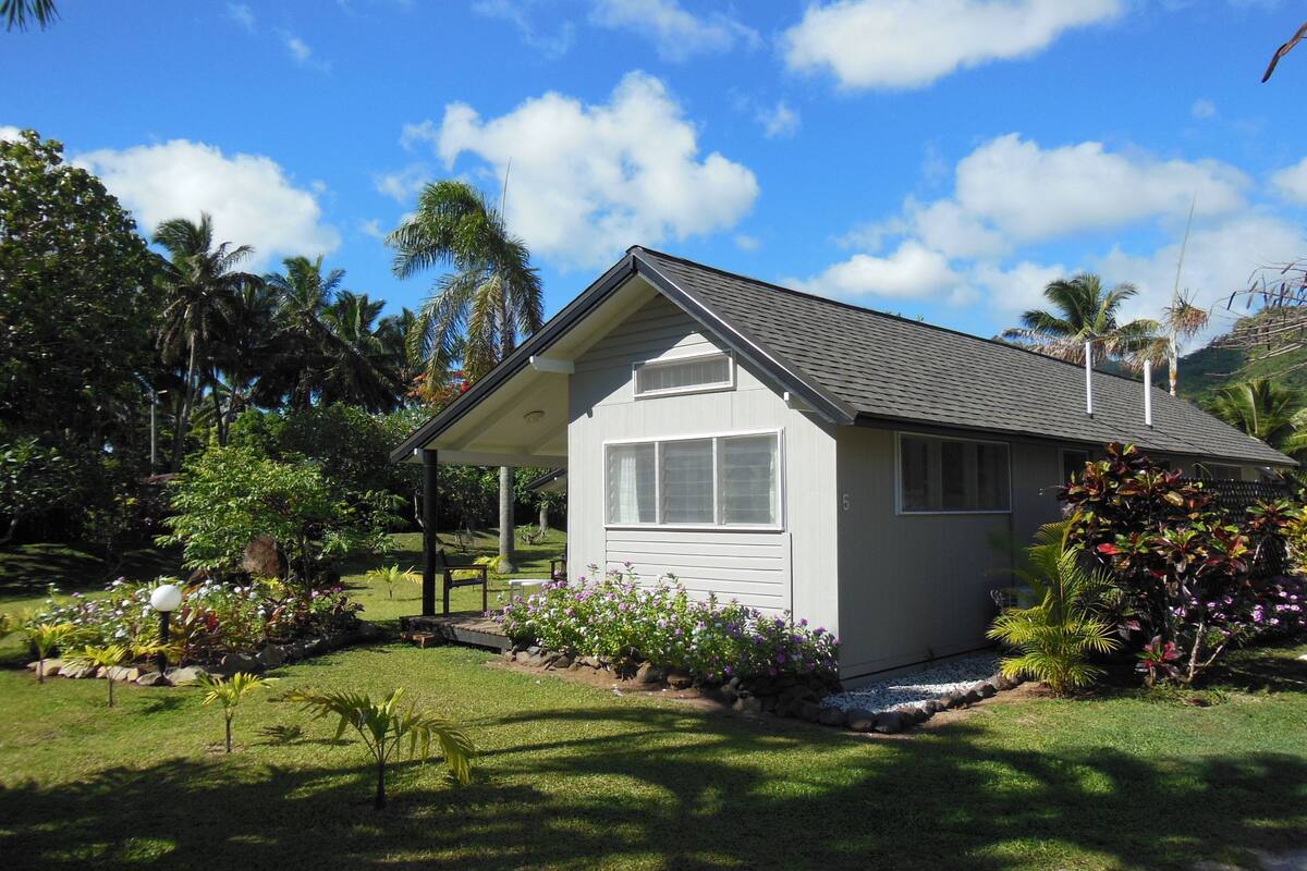 A single-story house with a grey roof and white walls, surrounded by lush greenery and palm trees.
