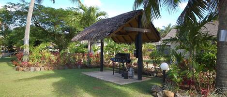 Un espacio al aire libre techado con una parrilla, rodeado de plantas tropicales y palmeras.