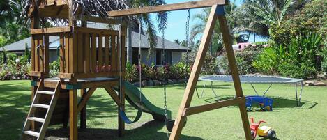 A wooden play structure with a slide and a swing set in a grassy yard with palm trees and a trampoline.
