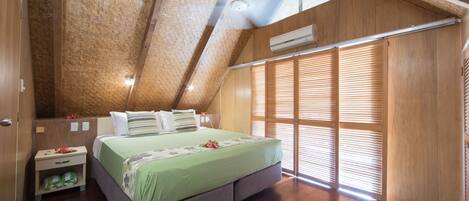 A bedroom with a sloped ceiling, a bed with a green bedspread, wooden walls, and a window with blinds.