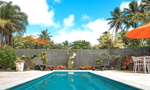 A swimming pool with two lounge chairs and an orange umbrella, surrounded by tropical plants and a wooden fence.