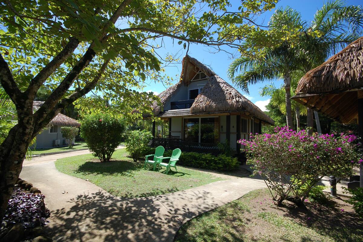 A thatched-roof house with a covered porch, surrounded by lush greenery and a clear sky.