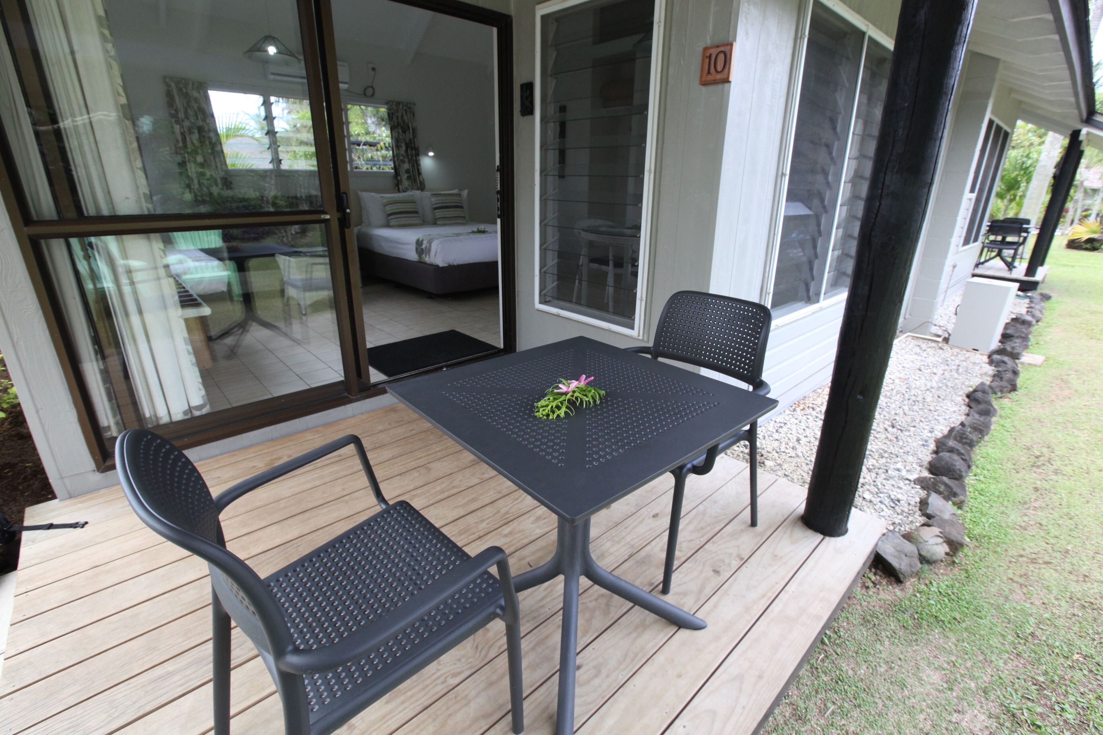Un patio couvert avec une table et deux chaises, une porte vitrée donnant sur une chambre, et une bordure en pierre le long du pourtour.
