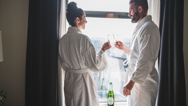 Two people in white bathrobes standing by a window, holding glasses.