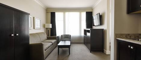 A hotel room with a dark wood wardrobe, a sofa, a coffee table, a TV, and a kitchen area with a countertop and cabinets.