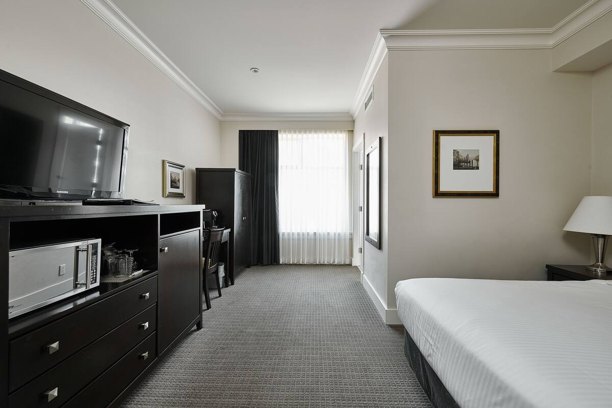 A hotel room with a bed, a television, a desk with a chair, and a window with curtains.