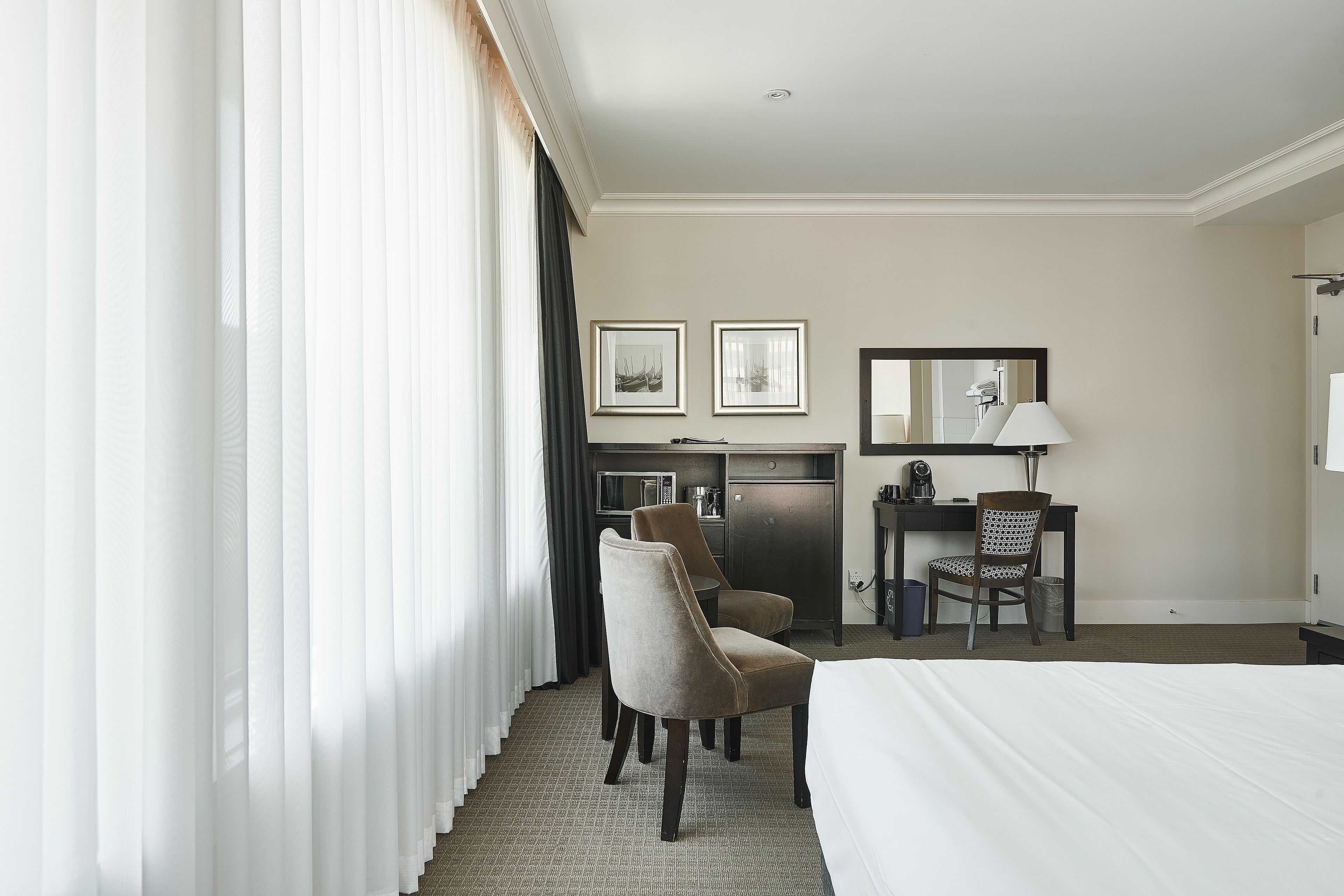 Hotelkamer met een bed, twee stoelen, een bureau met computer, een spiegel en twee ingelijste foto's aan de muur.