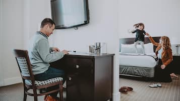 A hotel room with a desk, a bed, a TV, and a child playing on the bed.