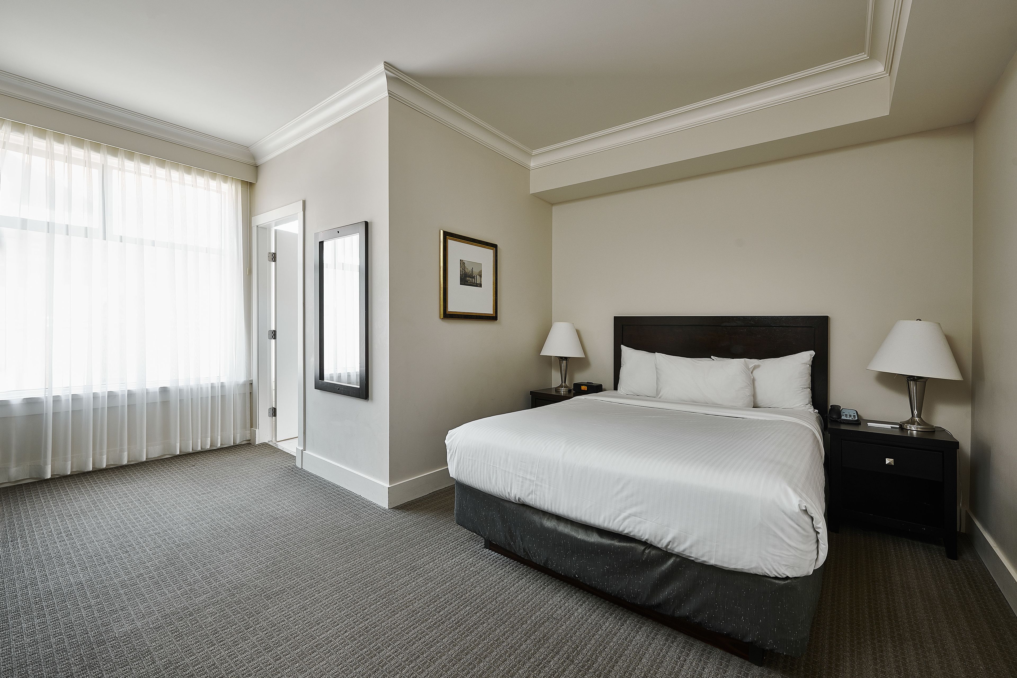 Een hotelkamer met een groot bed, twee nachtkastjes met lampen, een lijst met een foto aan de muur en een raam met dunne gordijnen.