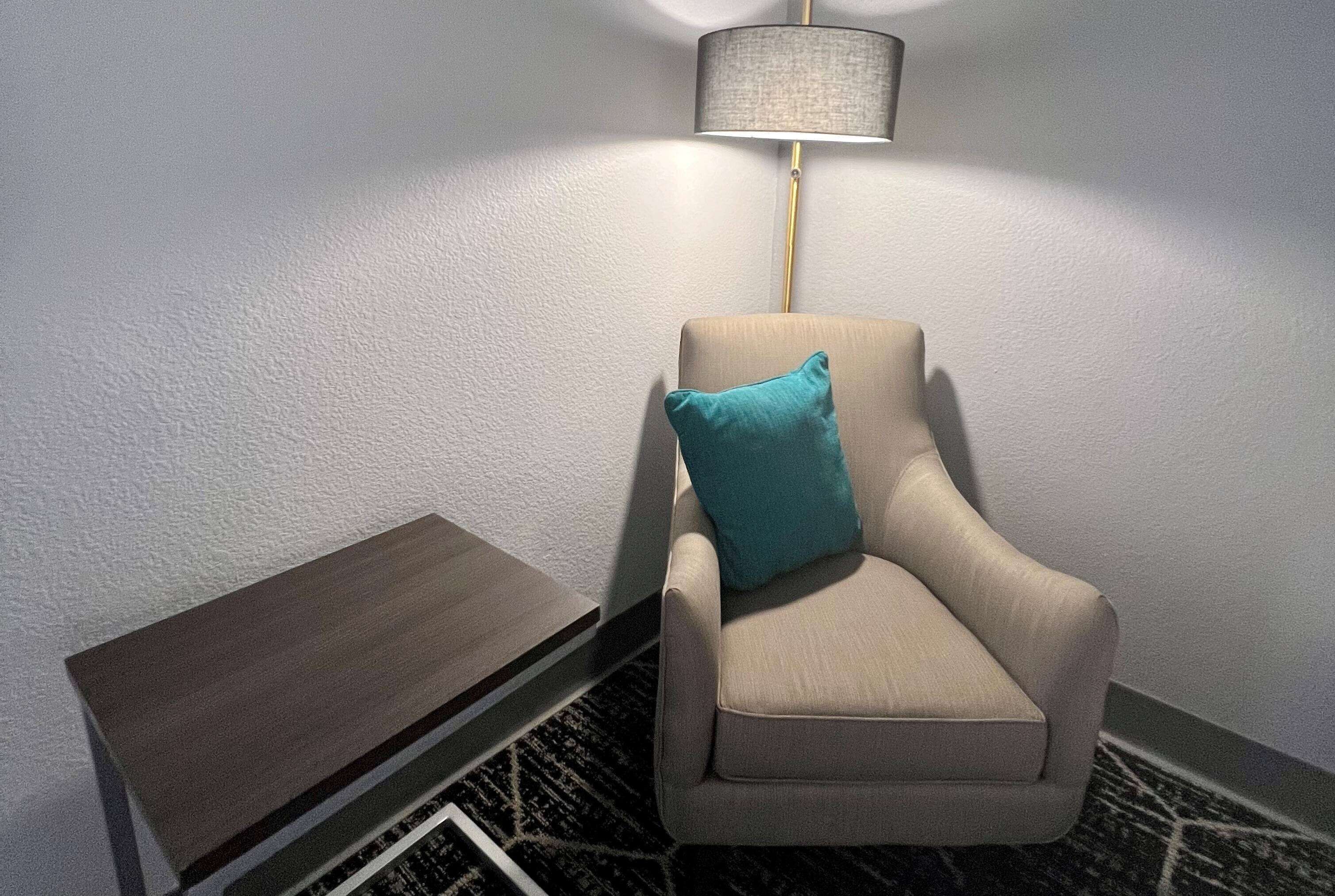 Een hoek van een kamer met een beige fauteuil, een blauwgroen kussen, een klein houten bijzettafeltje en een vloerlamp.