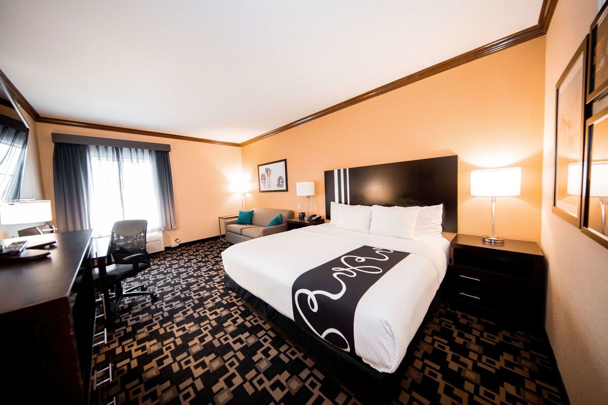 A hotel room with a large bed, a desk, a chair, a desk lamp, a nightstand, a framed picture on the wall, and a patterned carpet.