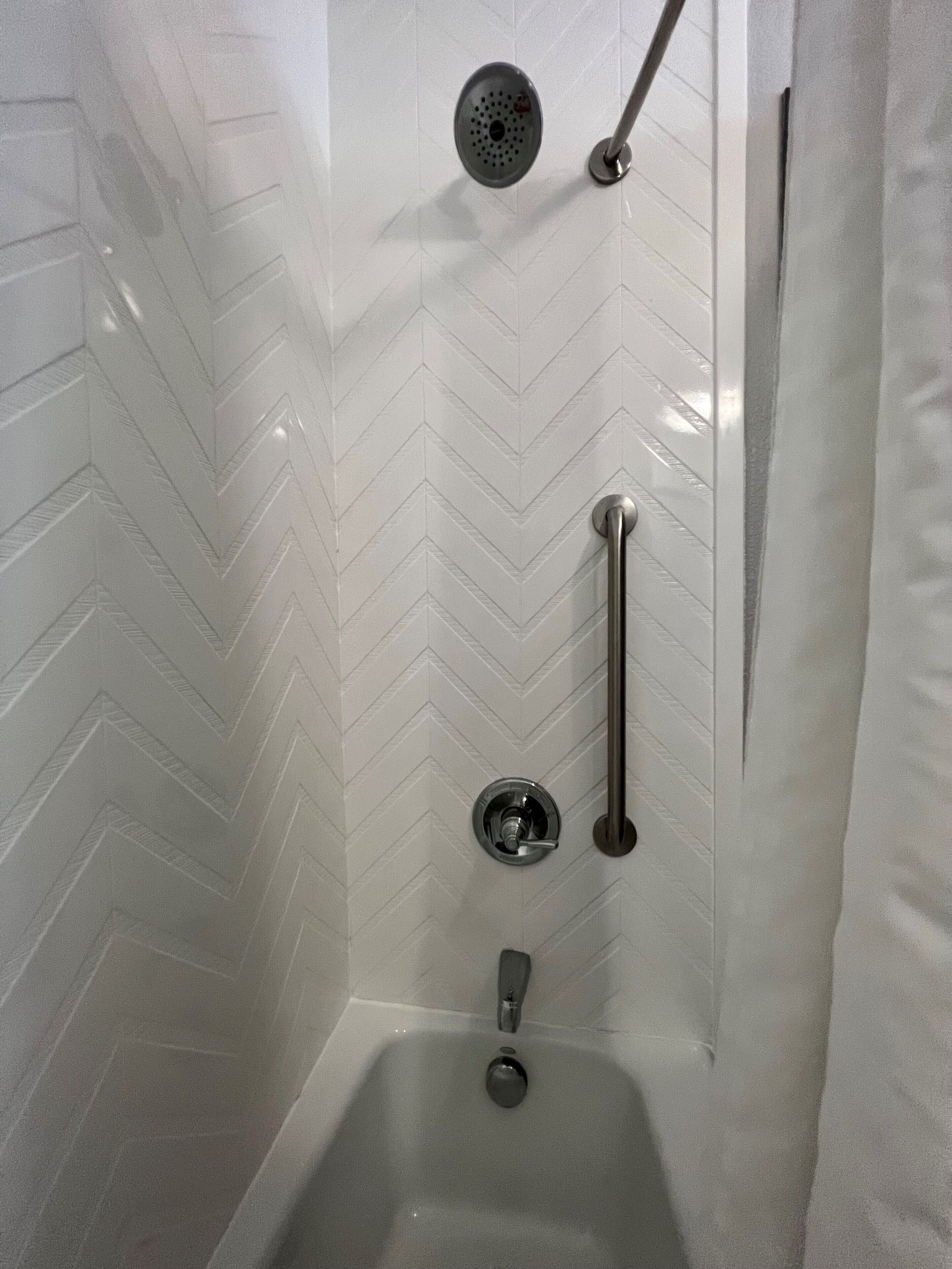 A shower with a white herringbone tile pattern, a handheld showerhead, a handheld shower control, and a floor-mounted control.