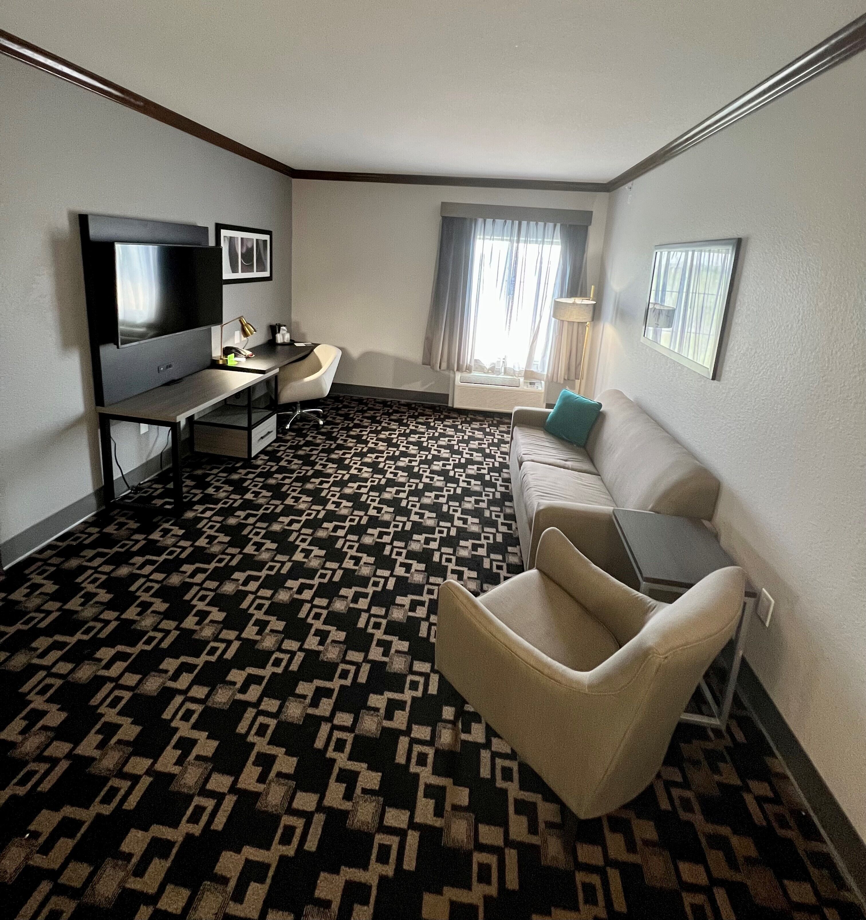 A hotel room with a flat-screen TV, a desk with a lamp, a sofa, and a patterned carpet.