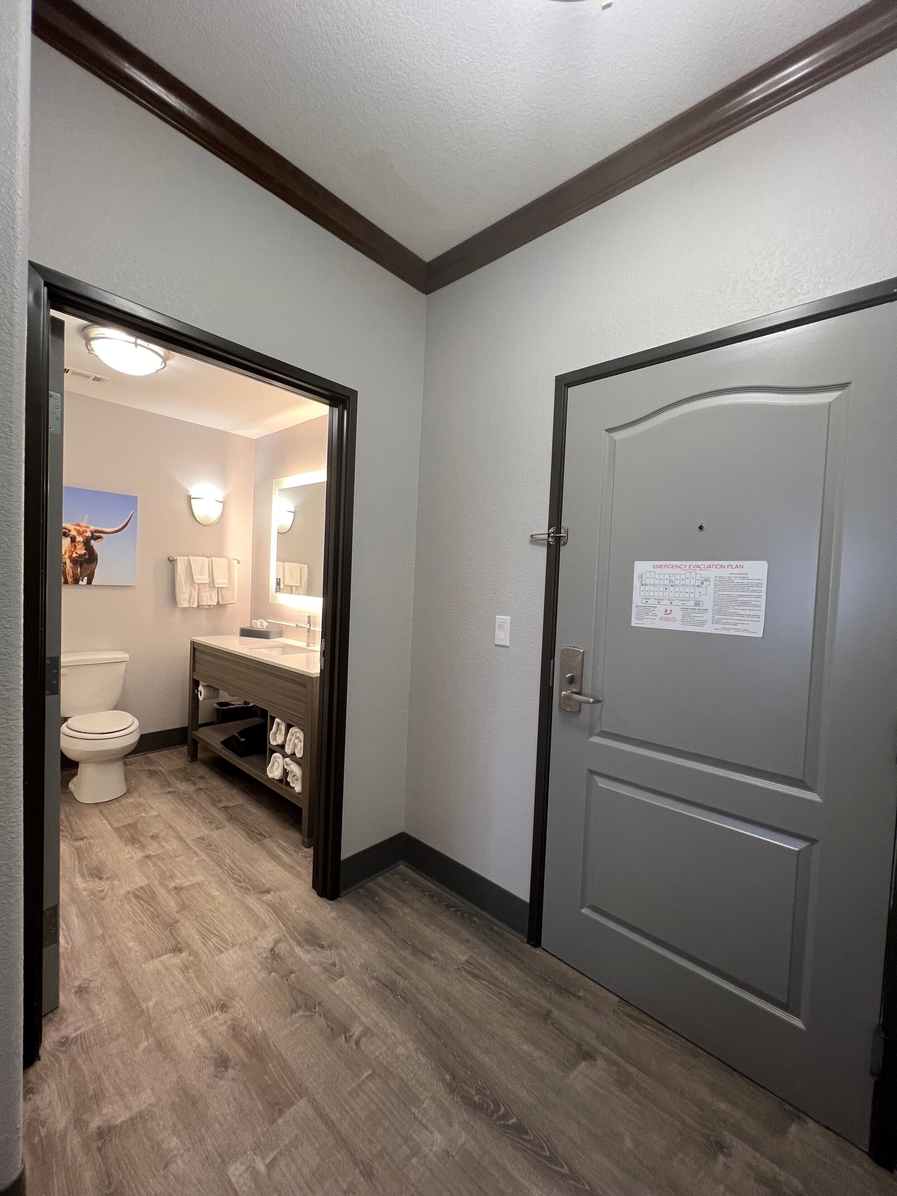 A hallway with a wooden floor, a bathroom with a toilet, sink, and towel rack, and a door with an emergency evacuation plan sign.