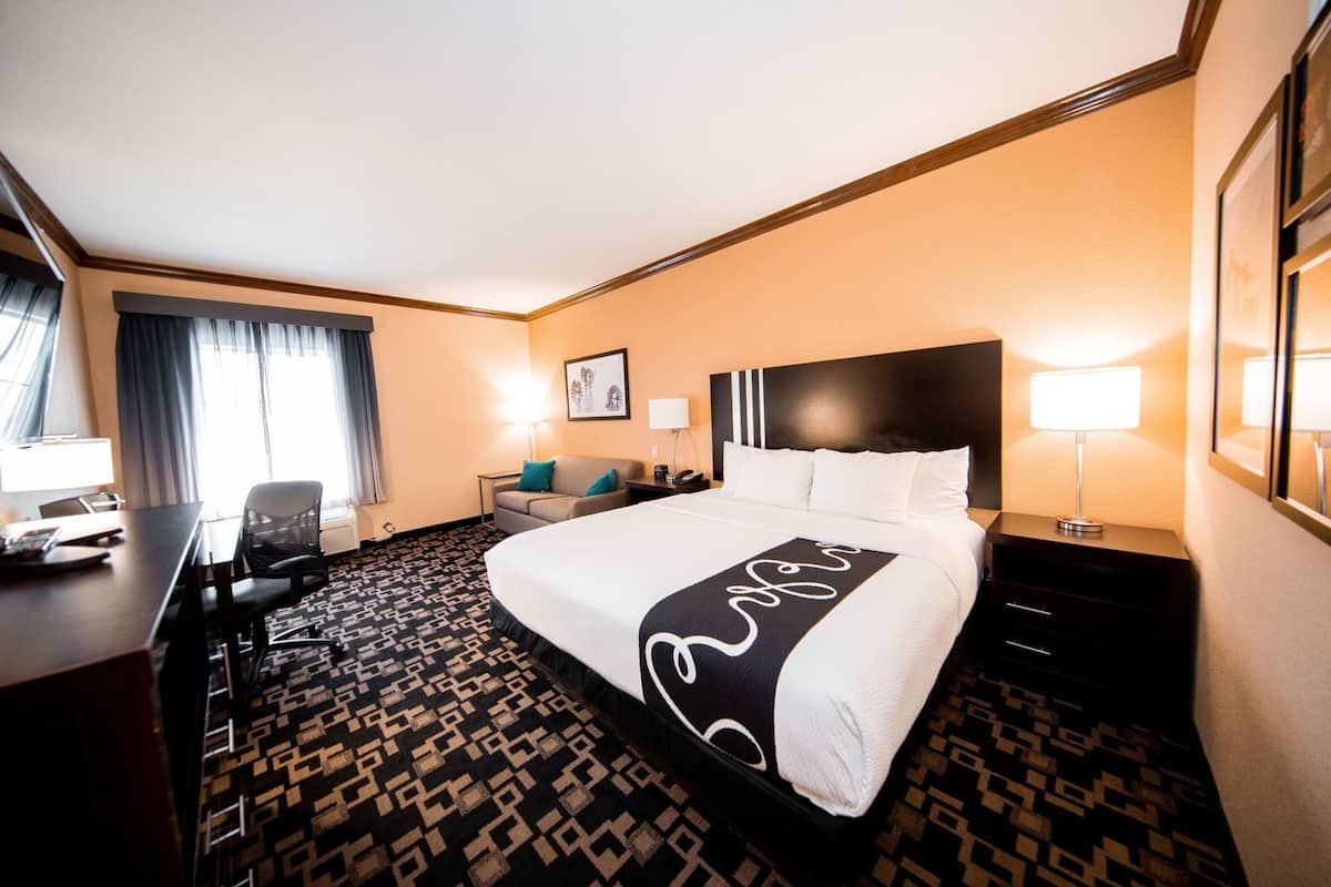 A hotel room with a large bed, a desk, a chair, a desk lamp, a nightstand, a framed picture on the wall, and a patterned carpet.