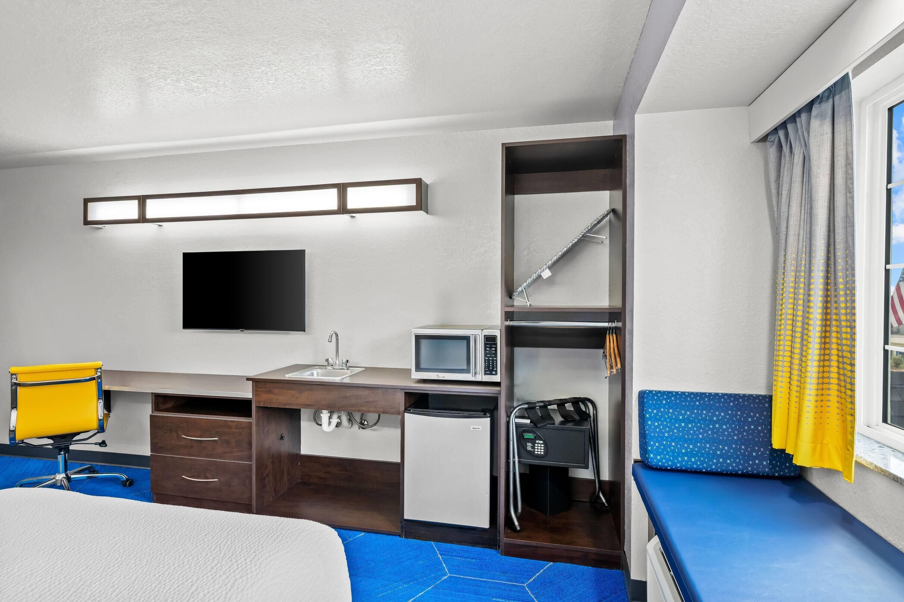 Habitación de hotel compacta con cama, escritorio con silla, microondas, refrigerador pequeño, televisor y ventana con cortinas.