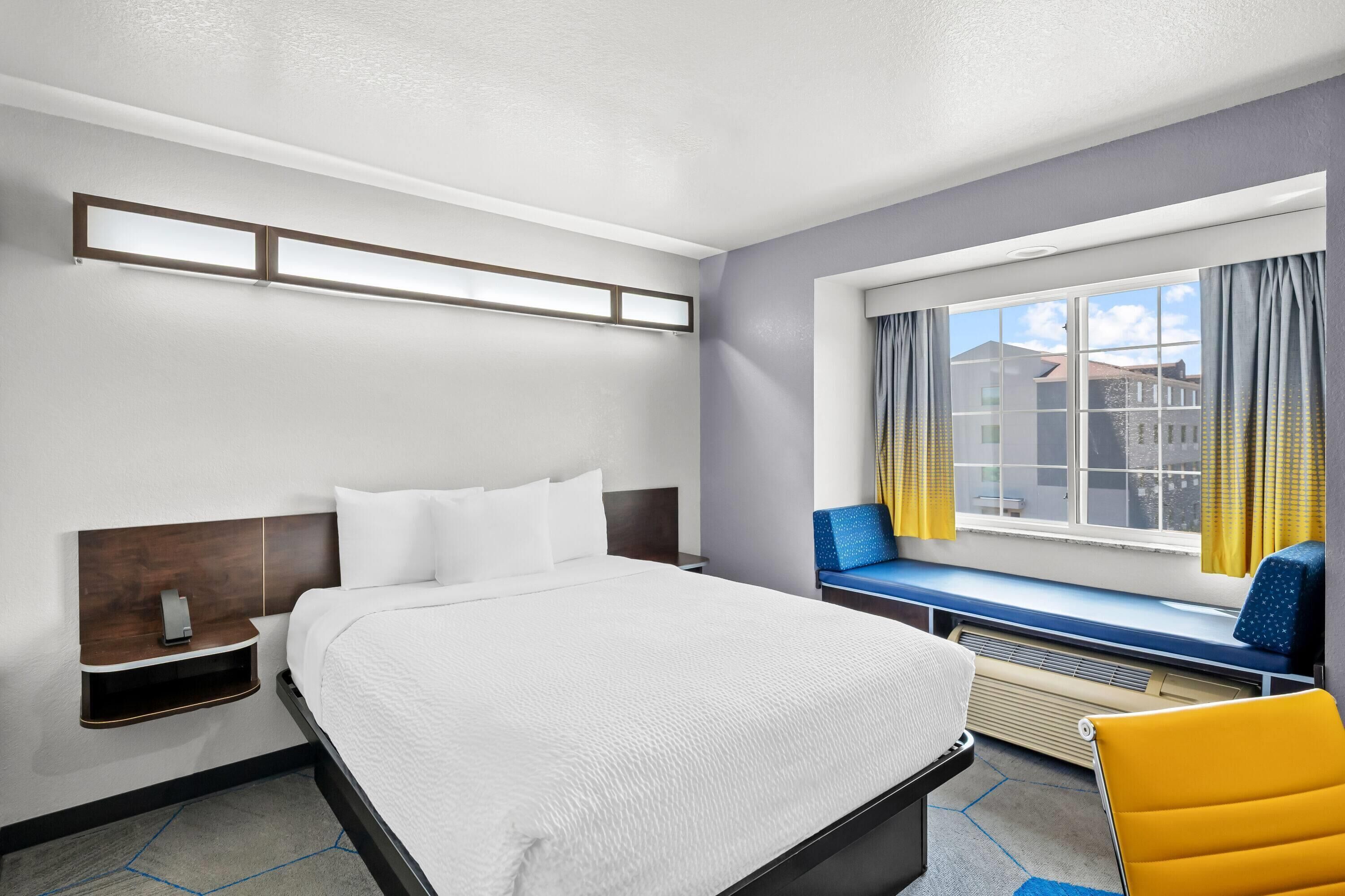 A hotel room with a large bed, a bedside table, a window with curtains, a bench, and a chair.