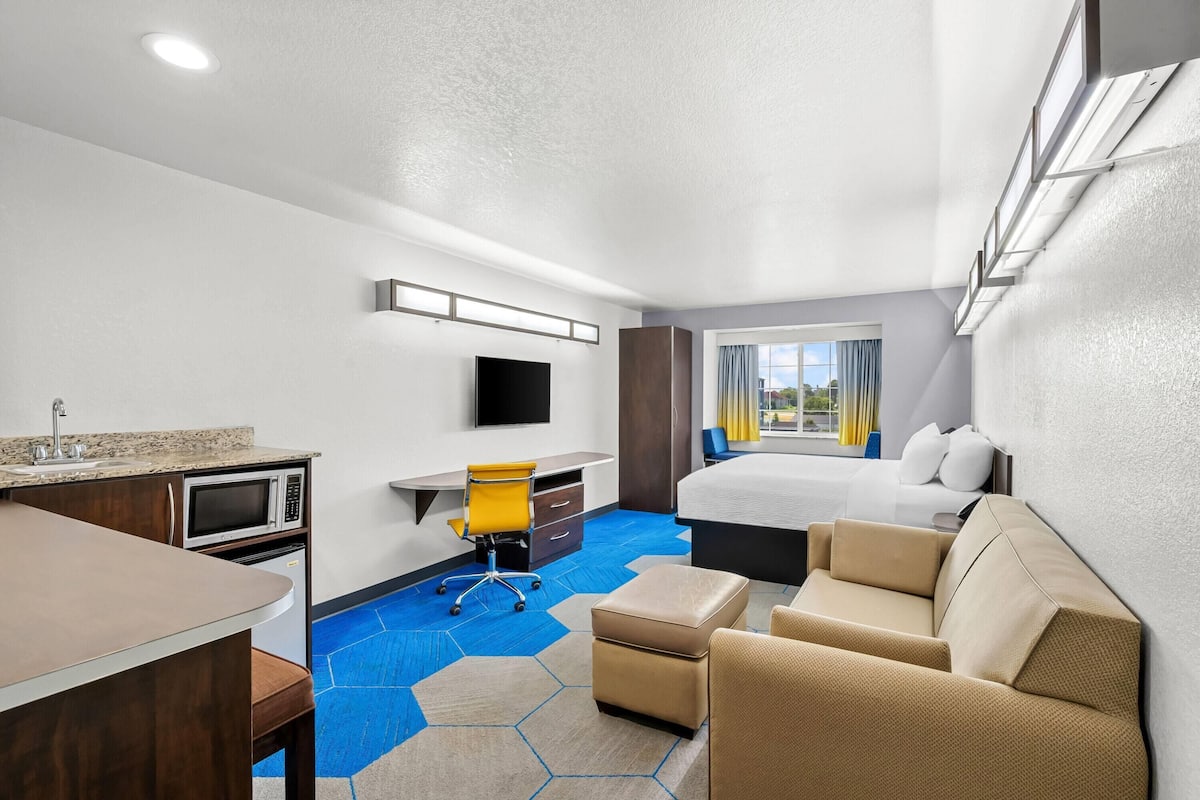 A hotel room with a bed, a desk with a chair, a kitchenette, and a large window with curtains.