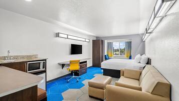 Quarto de hotel com cama, secretária e cadeira, cozinha compacta e uma janela grande com cortinas.