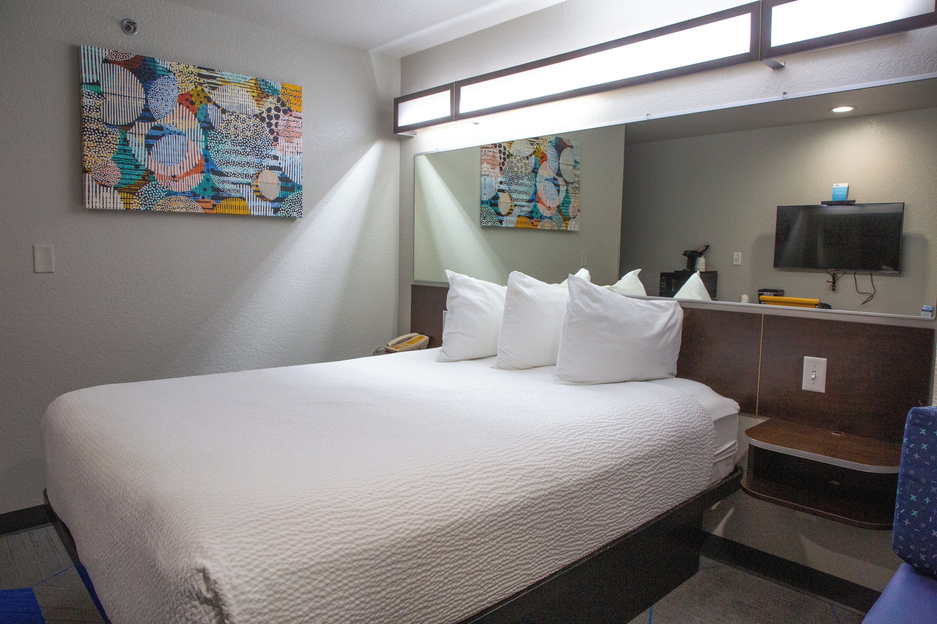 A hotel room with a large bed, a bedside table with a lamp, a TV mounted on the wall, and two paintings on the wall.