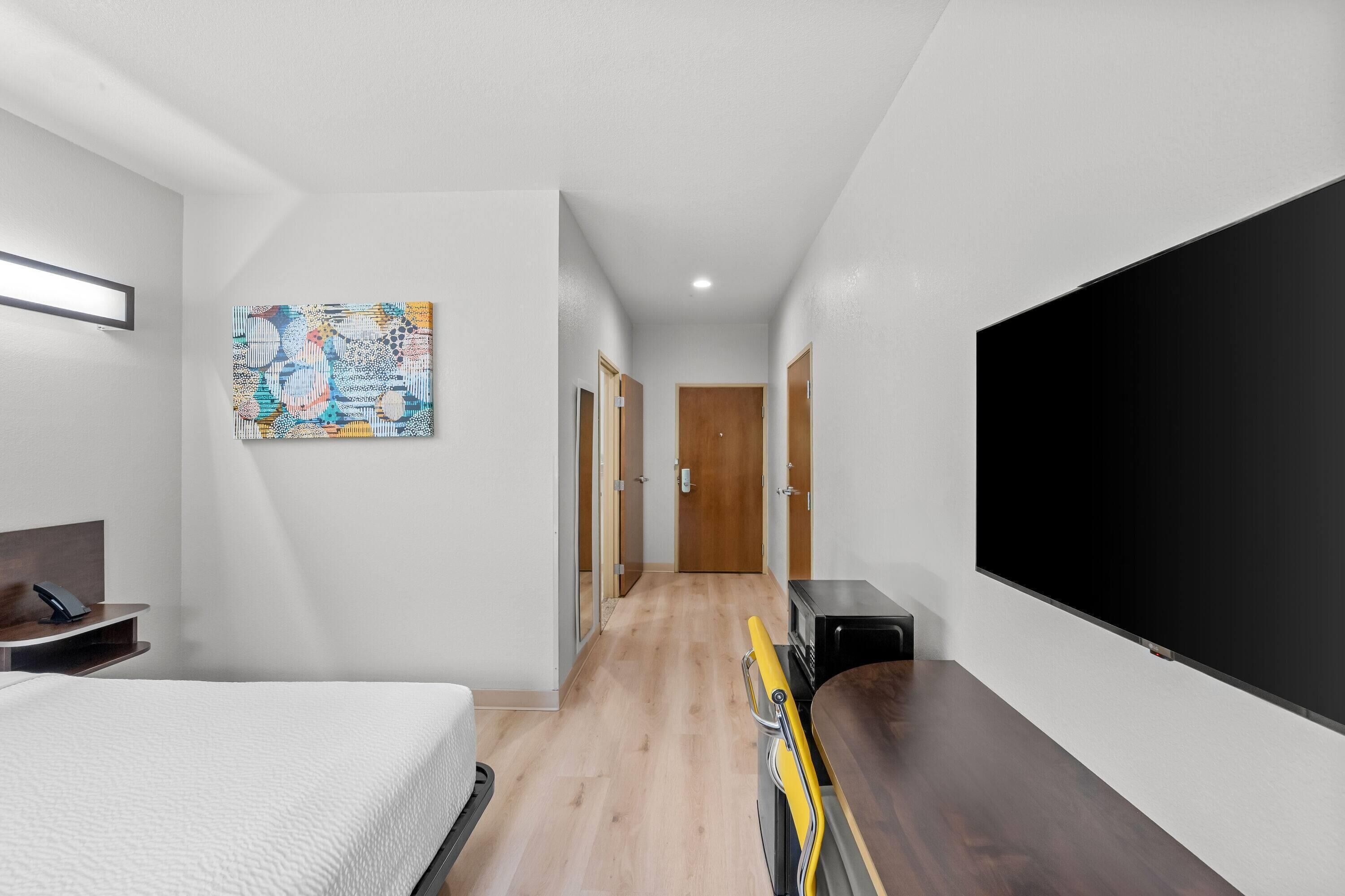 Quarto de hotel com cama, escrivaninha, televisão de tela plana e corredor que leva a uma porta.