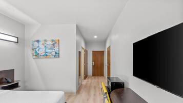 Quarto de hotel com cama, escrivaninha, televisão de tela plana e corredor que leva a uma porta.