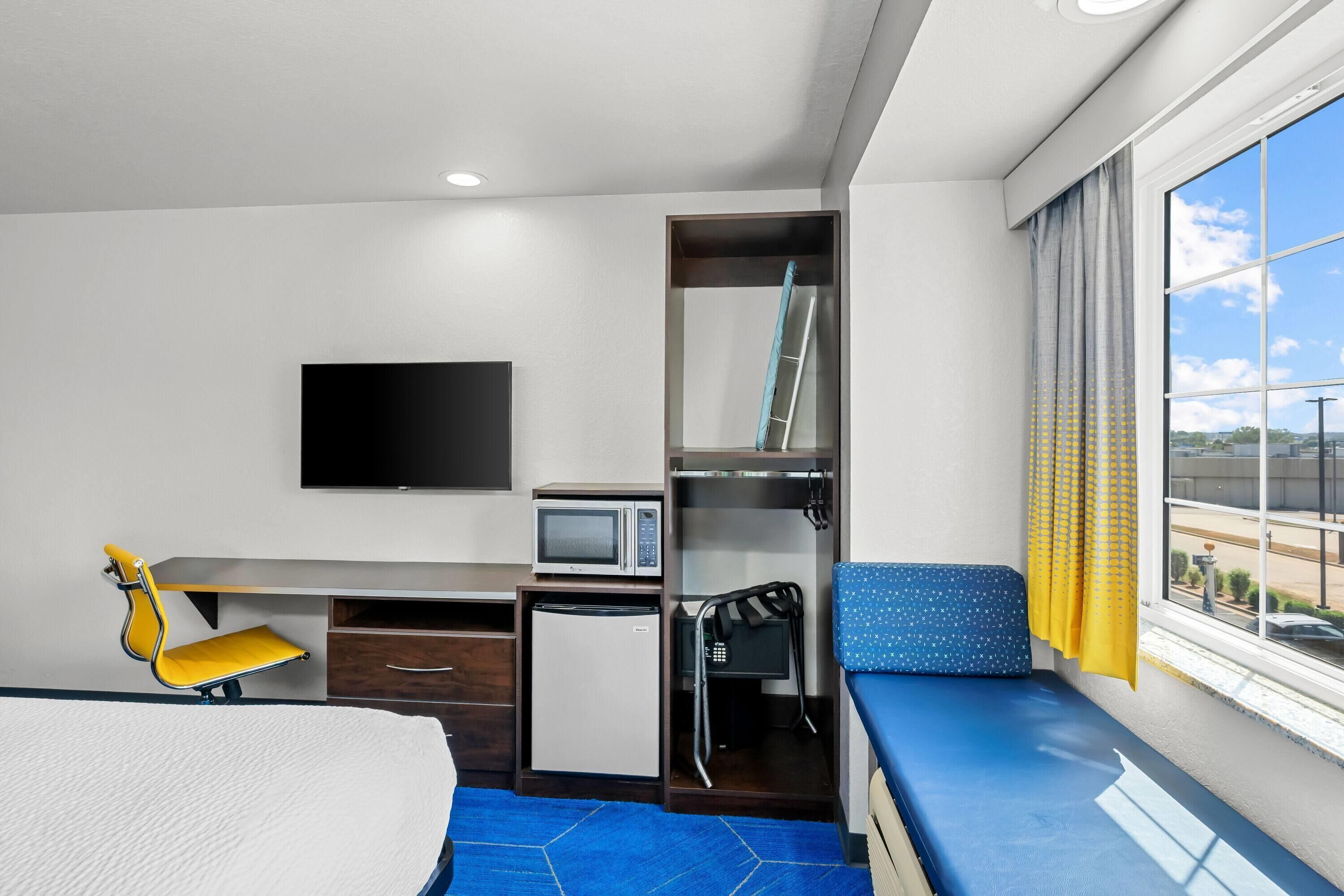 A compact hotel room with a bed, a desk with a computer, a microwave, a small fridge, a TV, and a window with curtains.