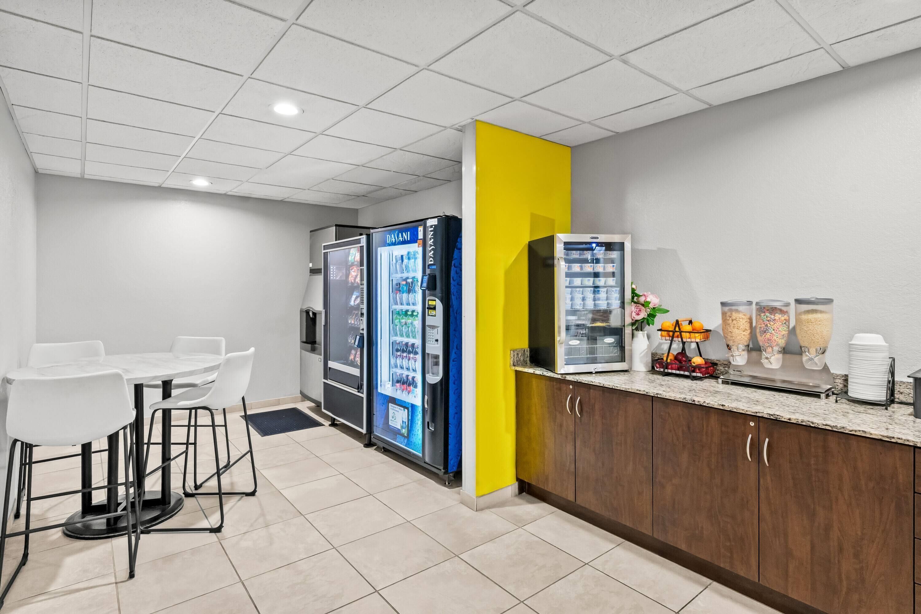 A modern kitchen area with a dining table and chairs, a refrigerator, a beverage dispenser, and a counter with food items.