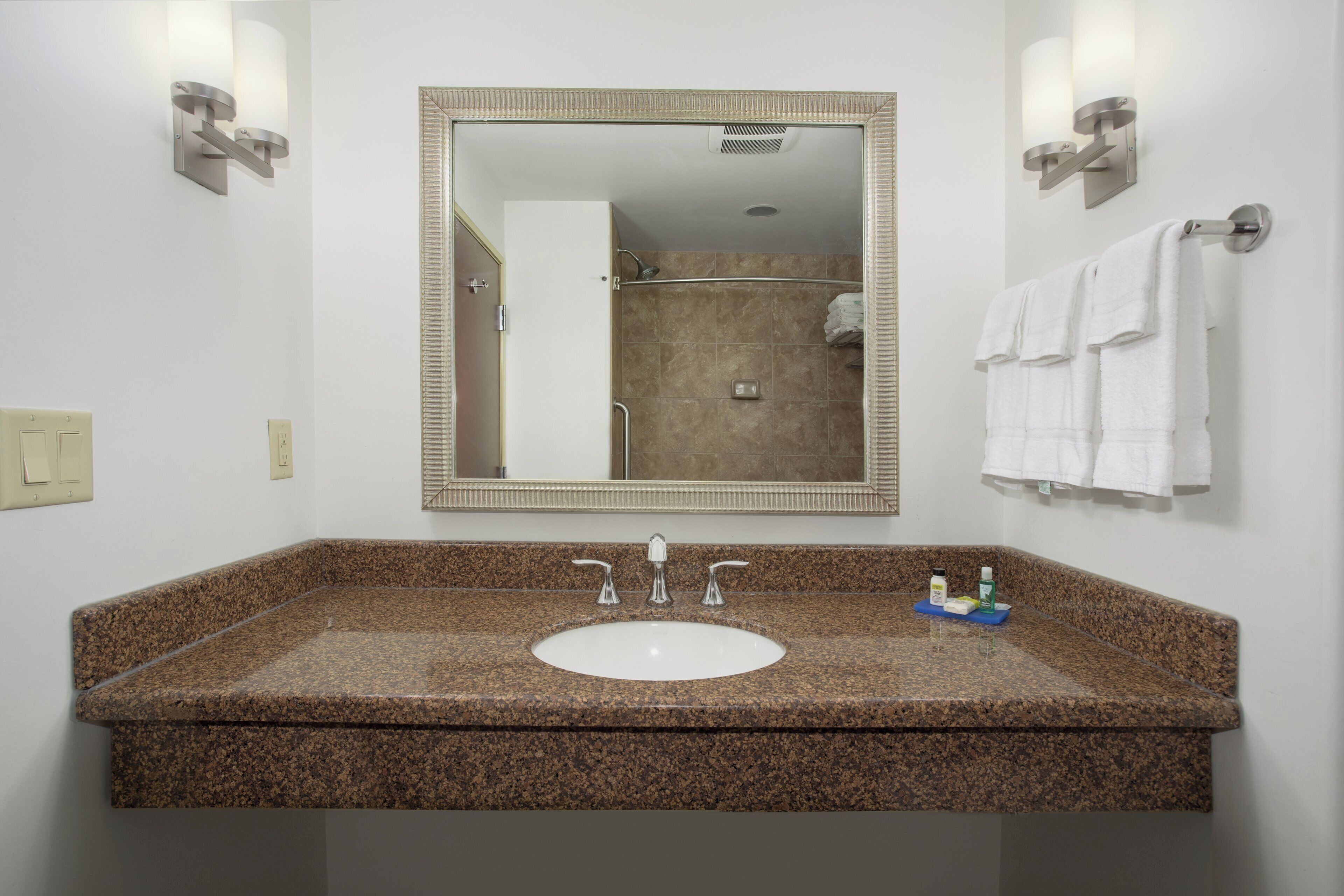 A bathroom with a granite countertop sink, a large mirror, and two wall-mounted light fixtures.