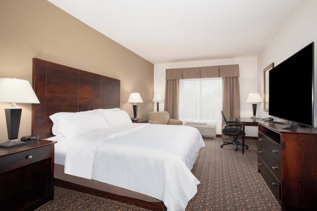 A hotel room with a large bed, two bedside tables with lamps, a desk with a chair, a flat-screen TV, and a large window with curtains.