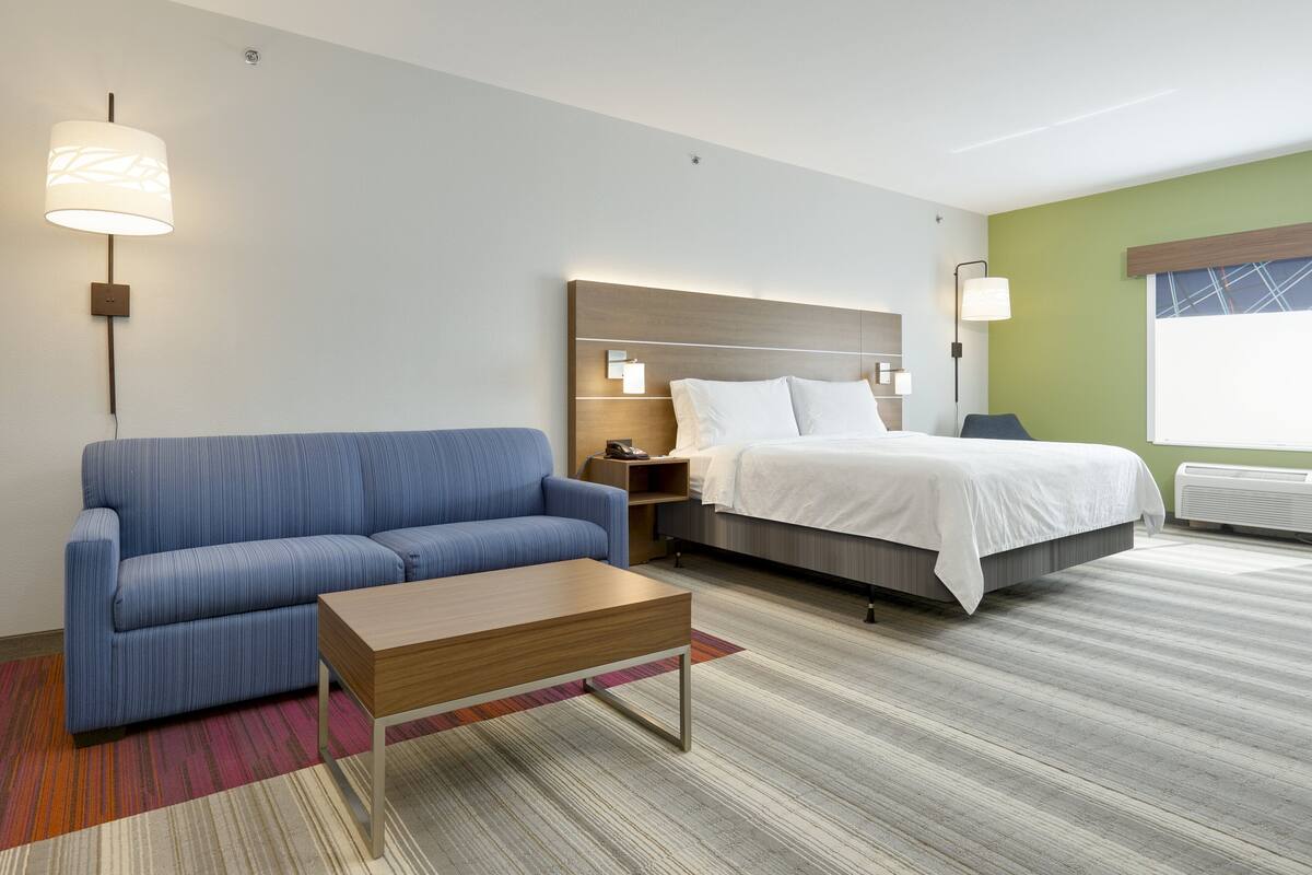 A hotel room with a blue sofa, a wooden coffee table, a bed with white bedding, and a nightstand with a lamp.