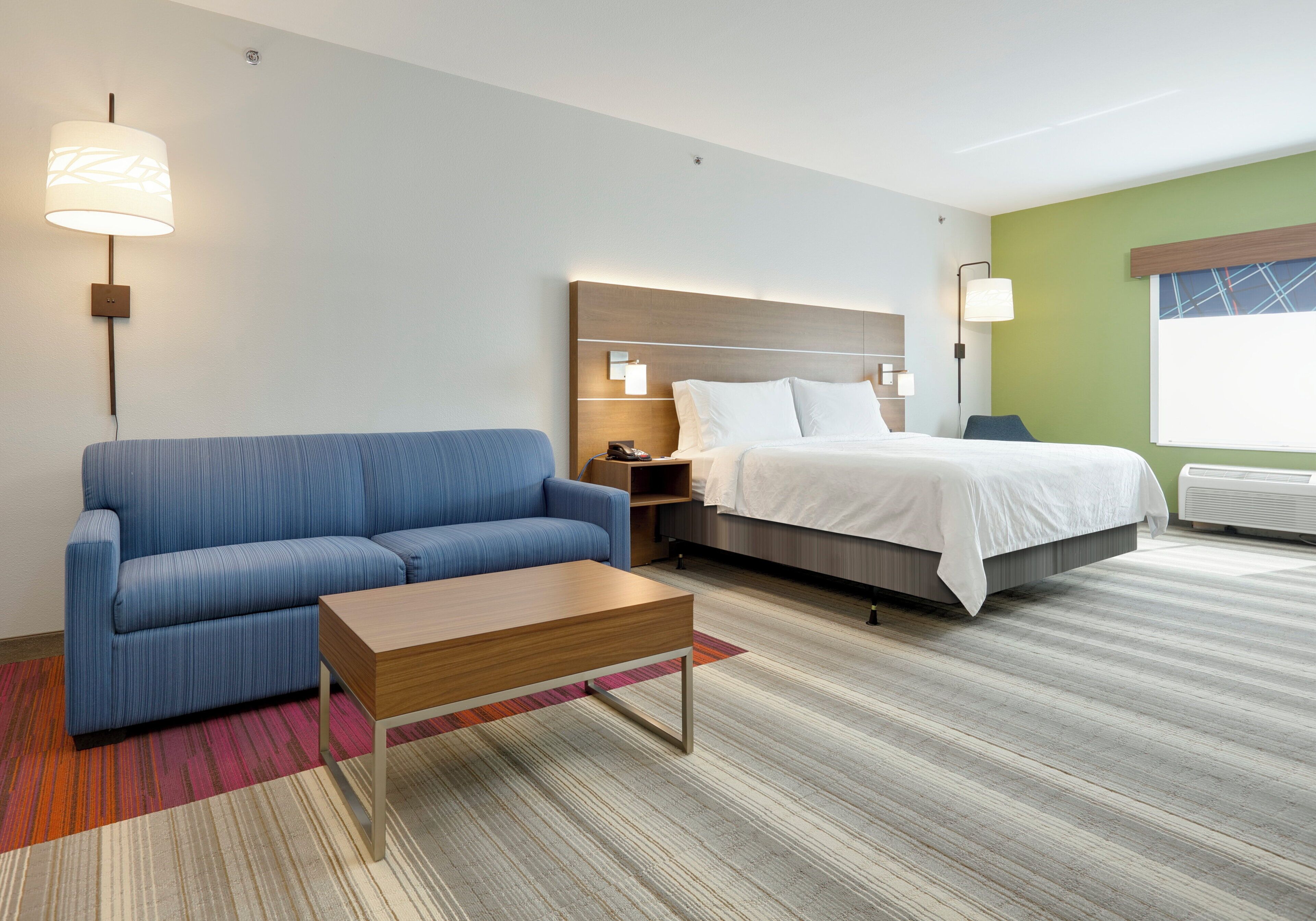 A hotel room with a blue sofa, a wooden side table, a bed with white linens, and a nightstand with a lamp.
