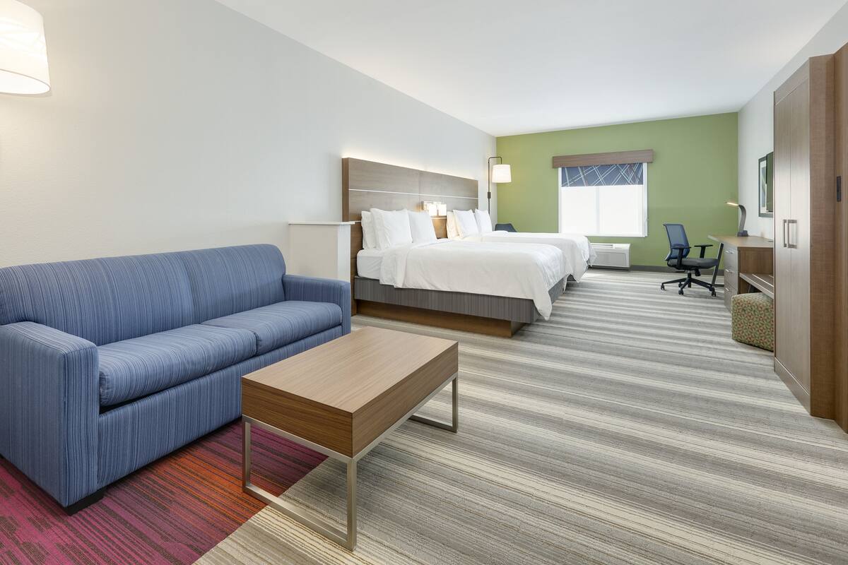 A hotel room with a blue sofa, a wooden coffee table, two beds with white linens, a desk with a chair, and a large wardrobe.