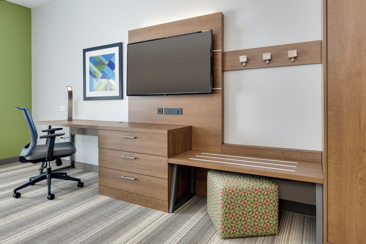 A modern office space with a wooden desk, a chair, a TV, and a patterned ottoman.
