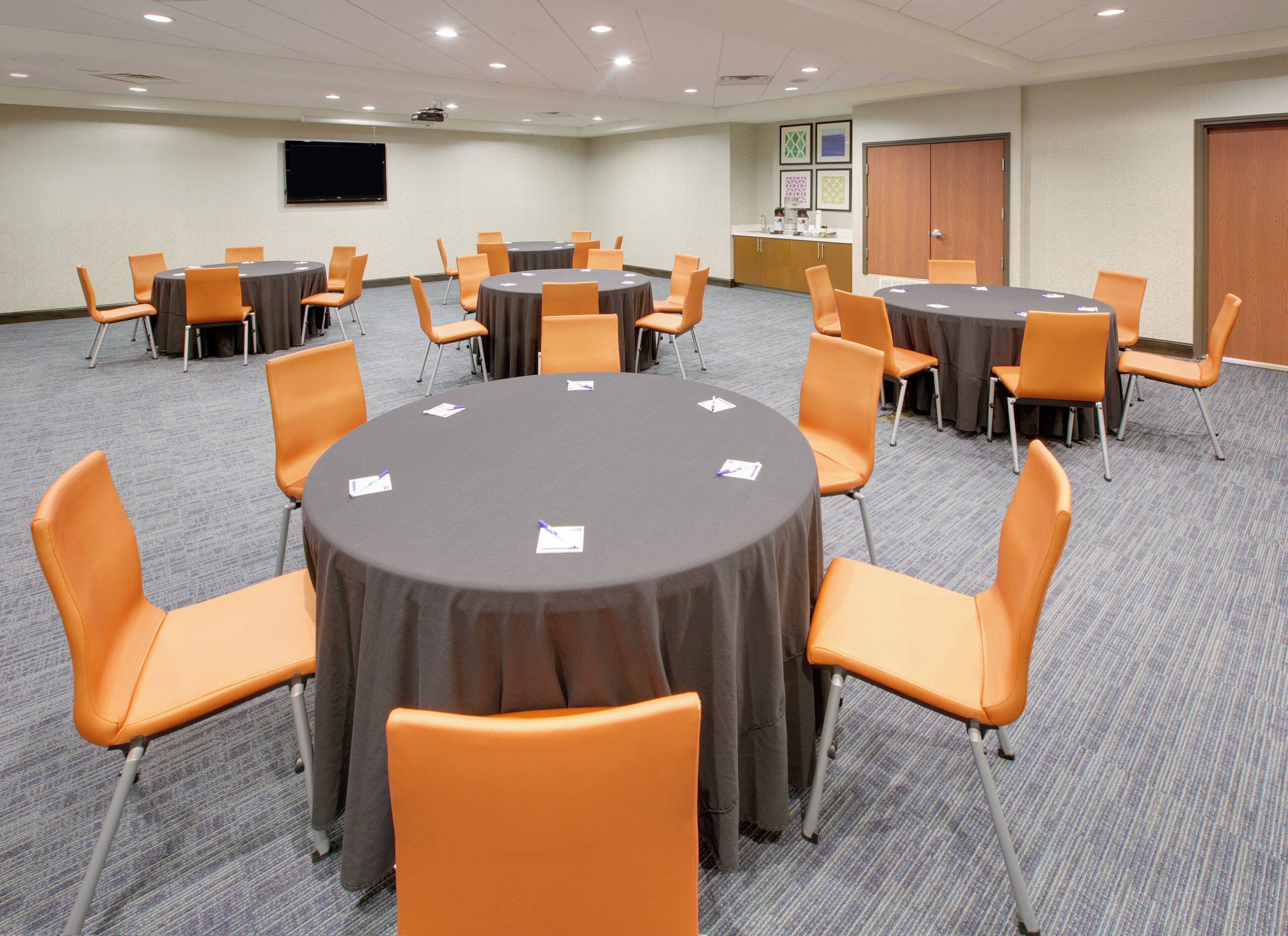 A conference room with multiple tables and chairs, a projector screen, and a kitchenette area.