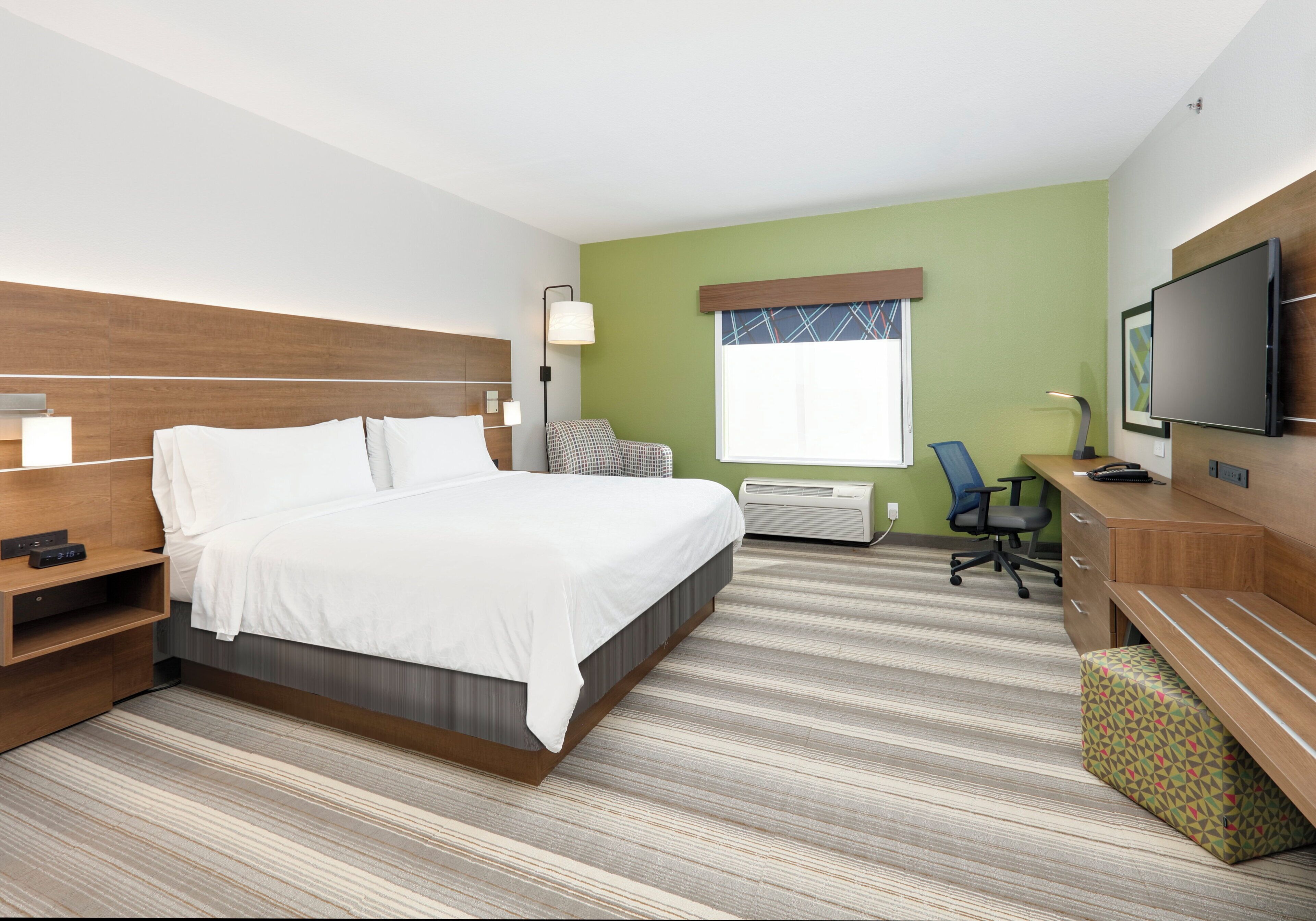 A hotel room with a bed, bedside table, lamp, TV, desk, chair, and air conditioning unit.